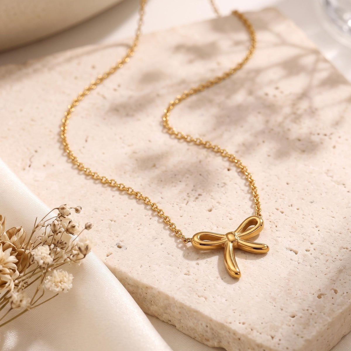Gold-tone bow pendant necklace displayed on a textured stone block with dried flowers.