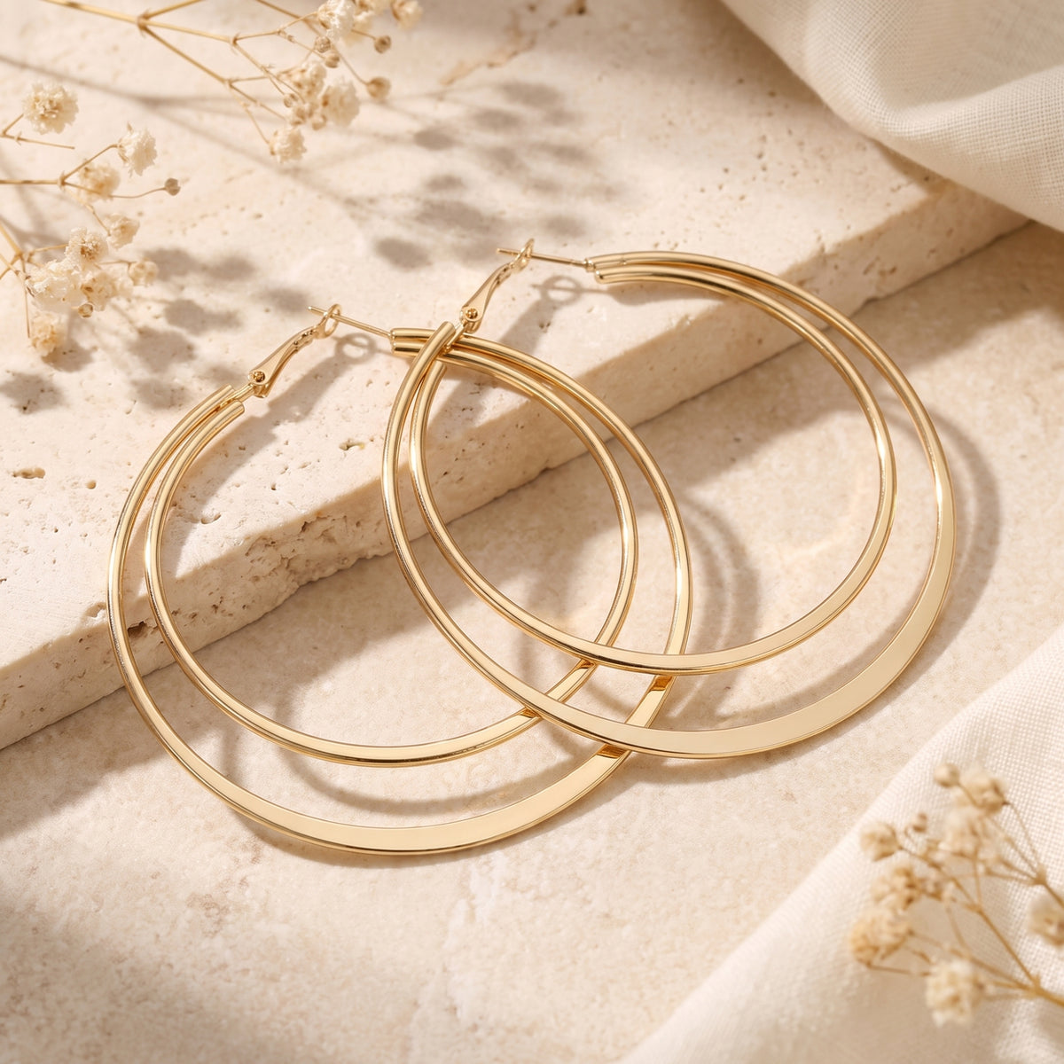 Gold-tone double hoop earrings resting on a pale stone slab beside dried flowers and light fabric.