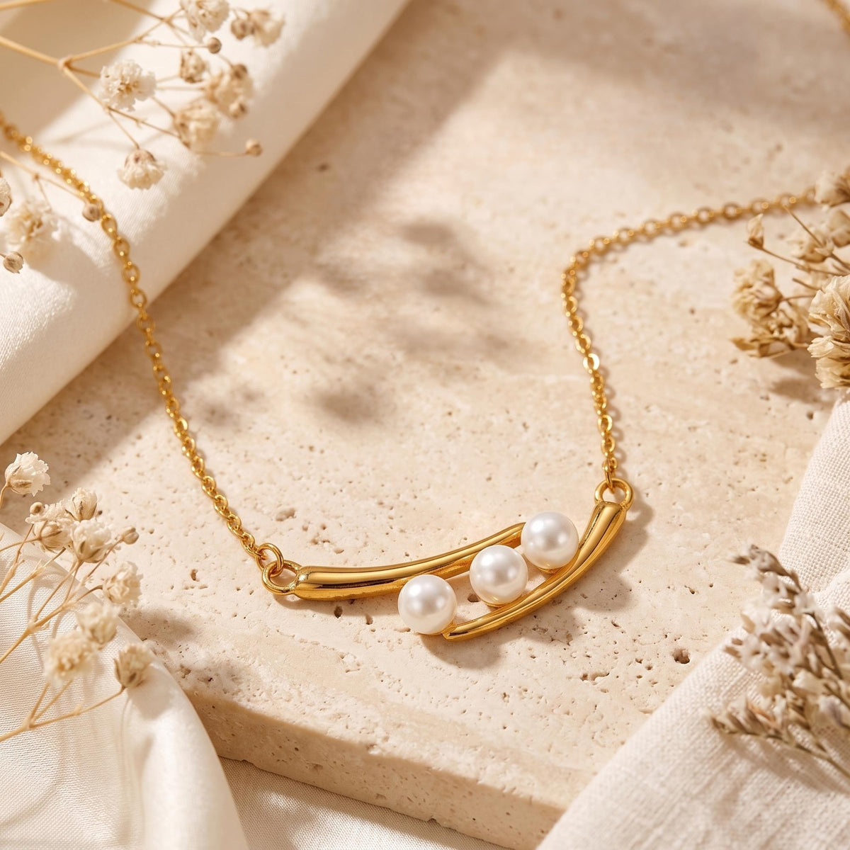 Gold-tone crescent necklace with three simulated pearls displayed on a pale stone slab with dried flowers.