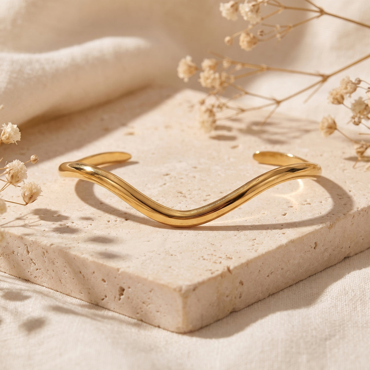 Gold-tone open wave cuff bracelet displayed horizontally on a cream stone slab with soft floral shadows.