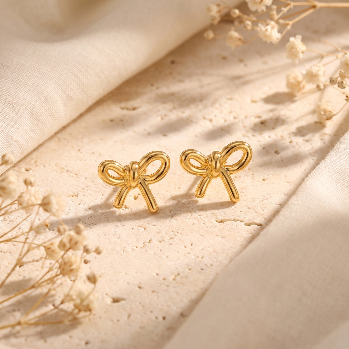 Pair of gold-toned bow drop earrings on a textured pale stone surface with dried flowers.