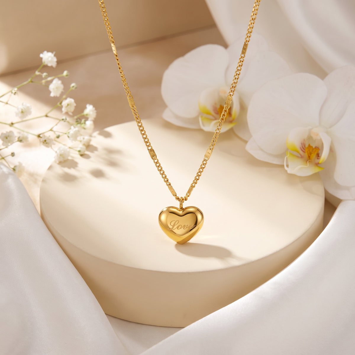 Gold-tone Love heart pendant necklace draped over a round beige pedestal with white orchids.