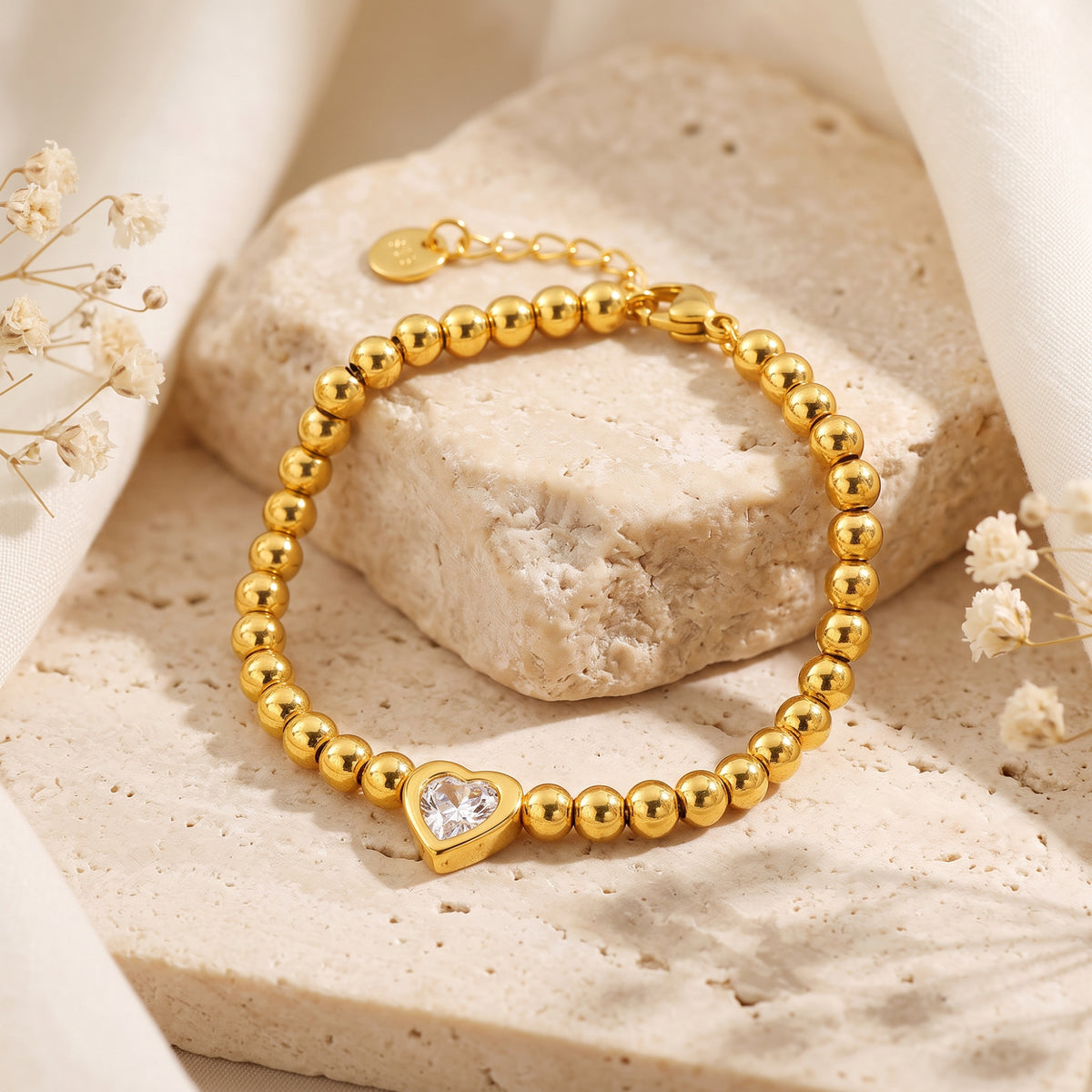 Gold-tone beaded bracelet with a bezel-set heart stone displayed in a loop on a textured stone surface.