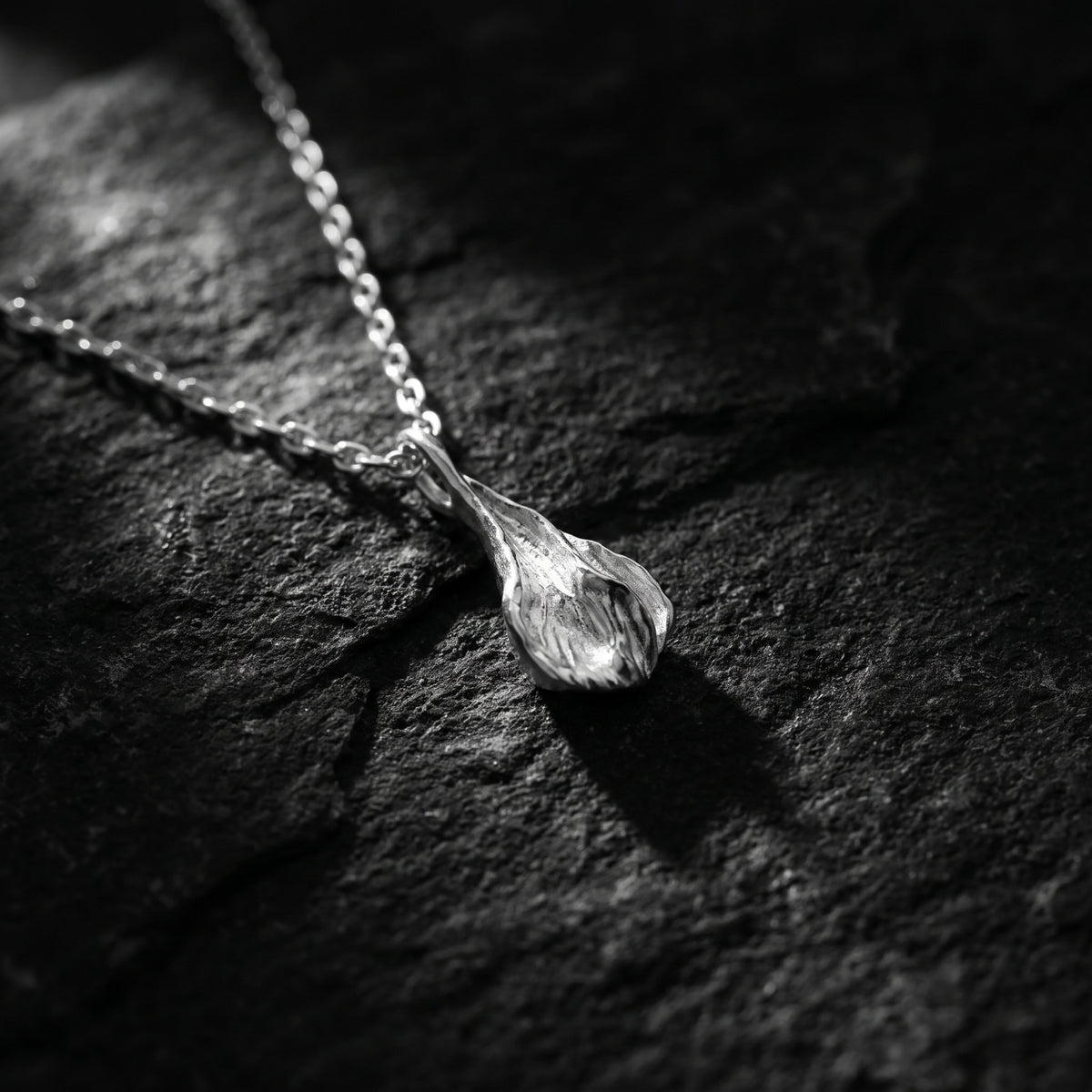 Close-up of the silver sculpted petal pendant resting diagonally on dark textured rock.