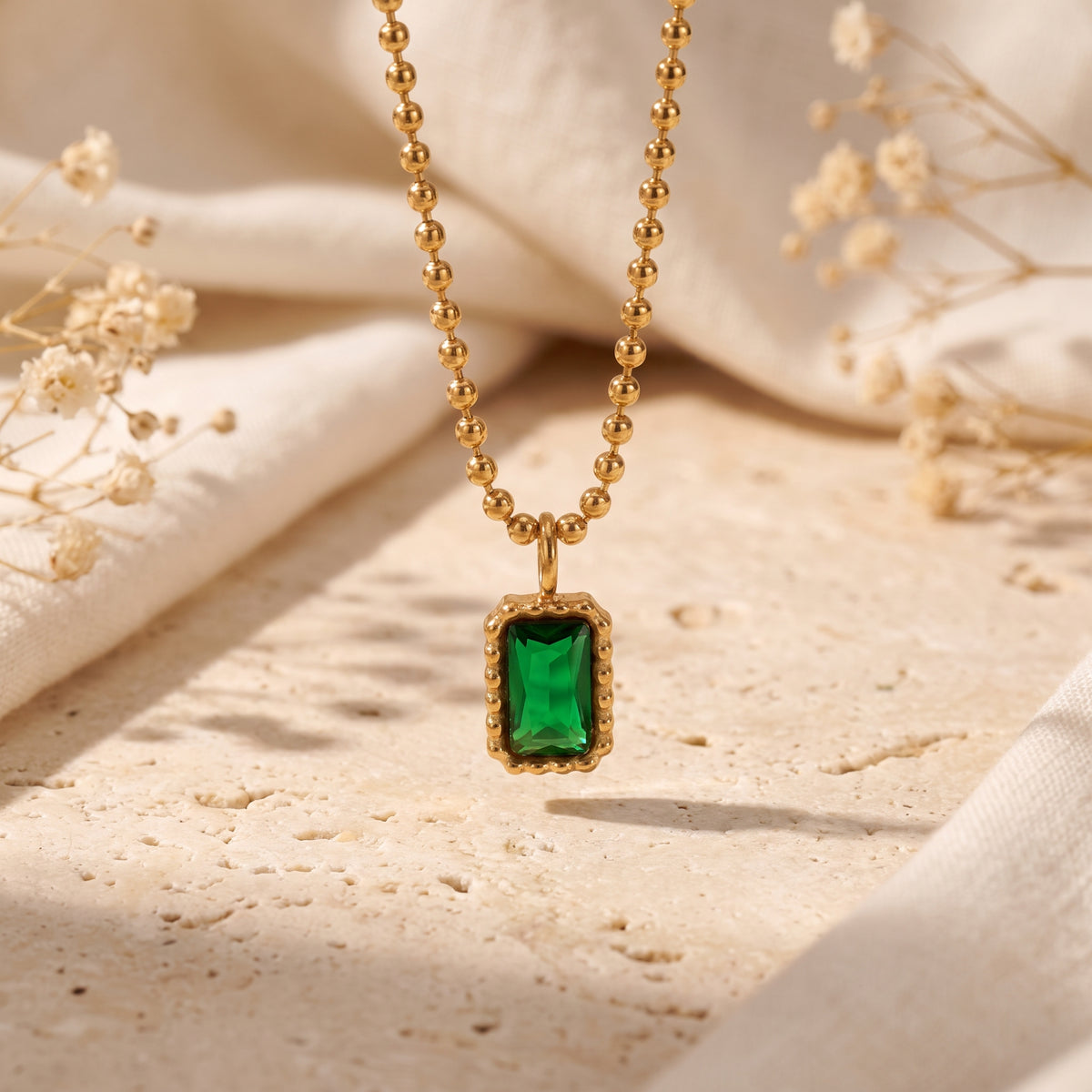Close-up of a green emerald-cut CZ pendant framed in a beaded gold-tone setting on a ball chain.