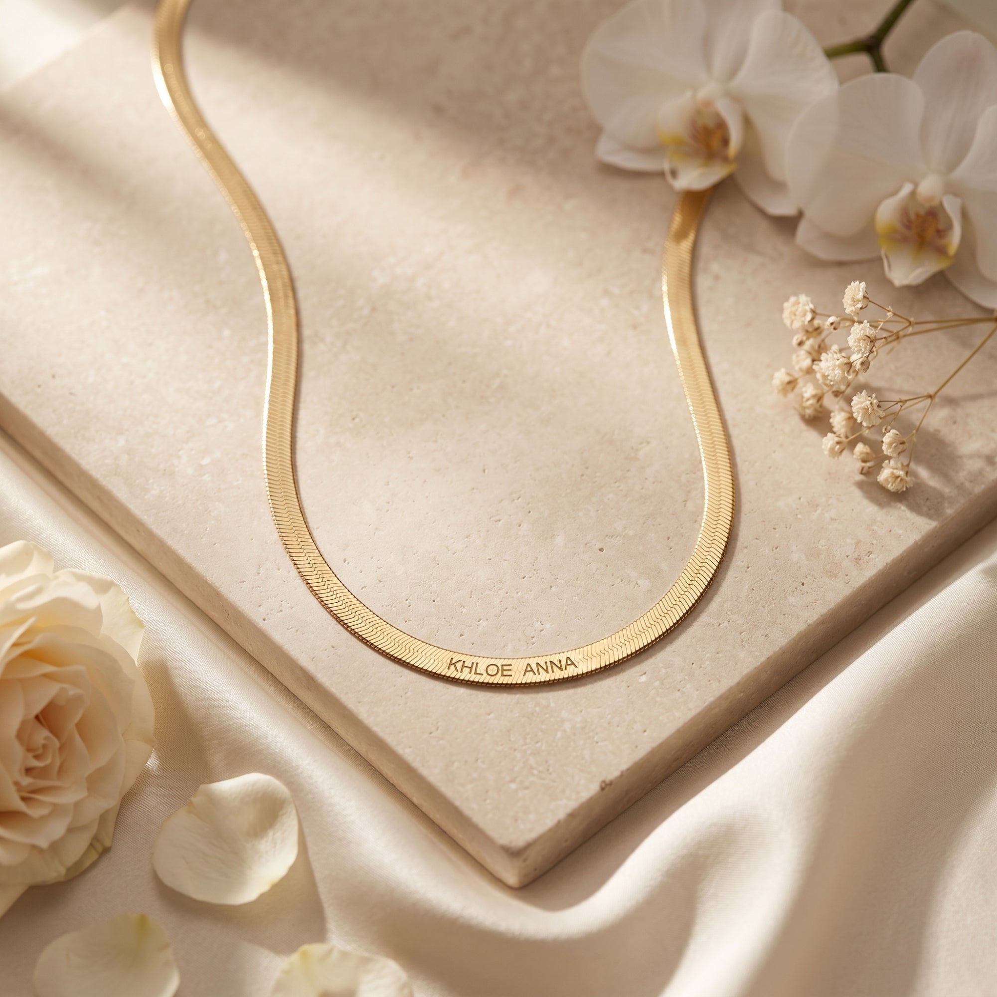 Custom 18K gold finish herringbone name necklace on stone with white flowers.