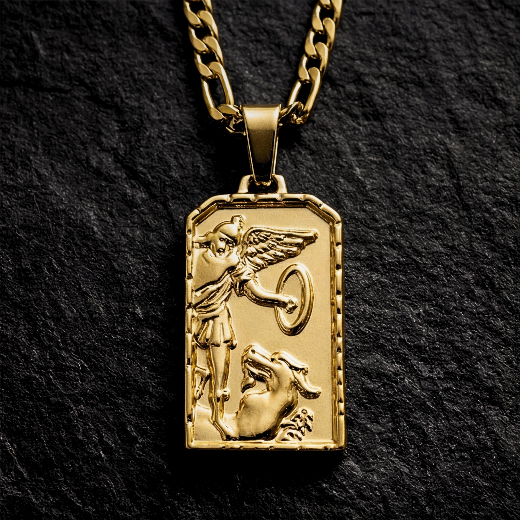 Gold stainless steel St. Michael Archangel pendant on a Figaro chain, displayed on a dark textured stone surface.