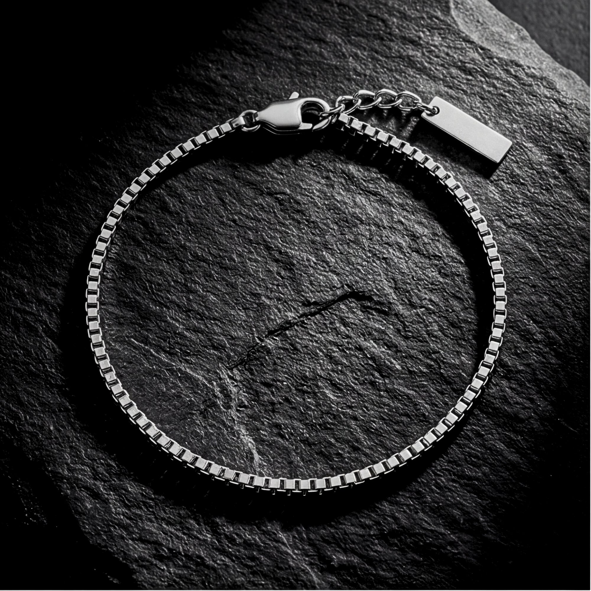 Silver stainless steel box chain bracelet with lobster clasp on a dark textured stone background.