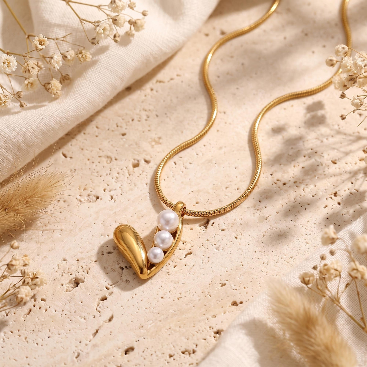 Flat lay of the gold-tone three-pearl pod pendant necklace on a beige stone surface with dried flowers.