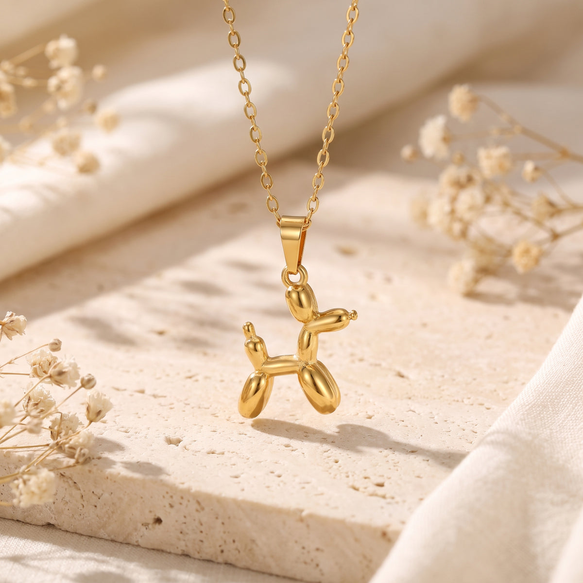 Gold-tone balloon dog pendant hanging suspended over a beige textured slab with dried flowers.
