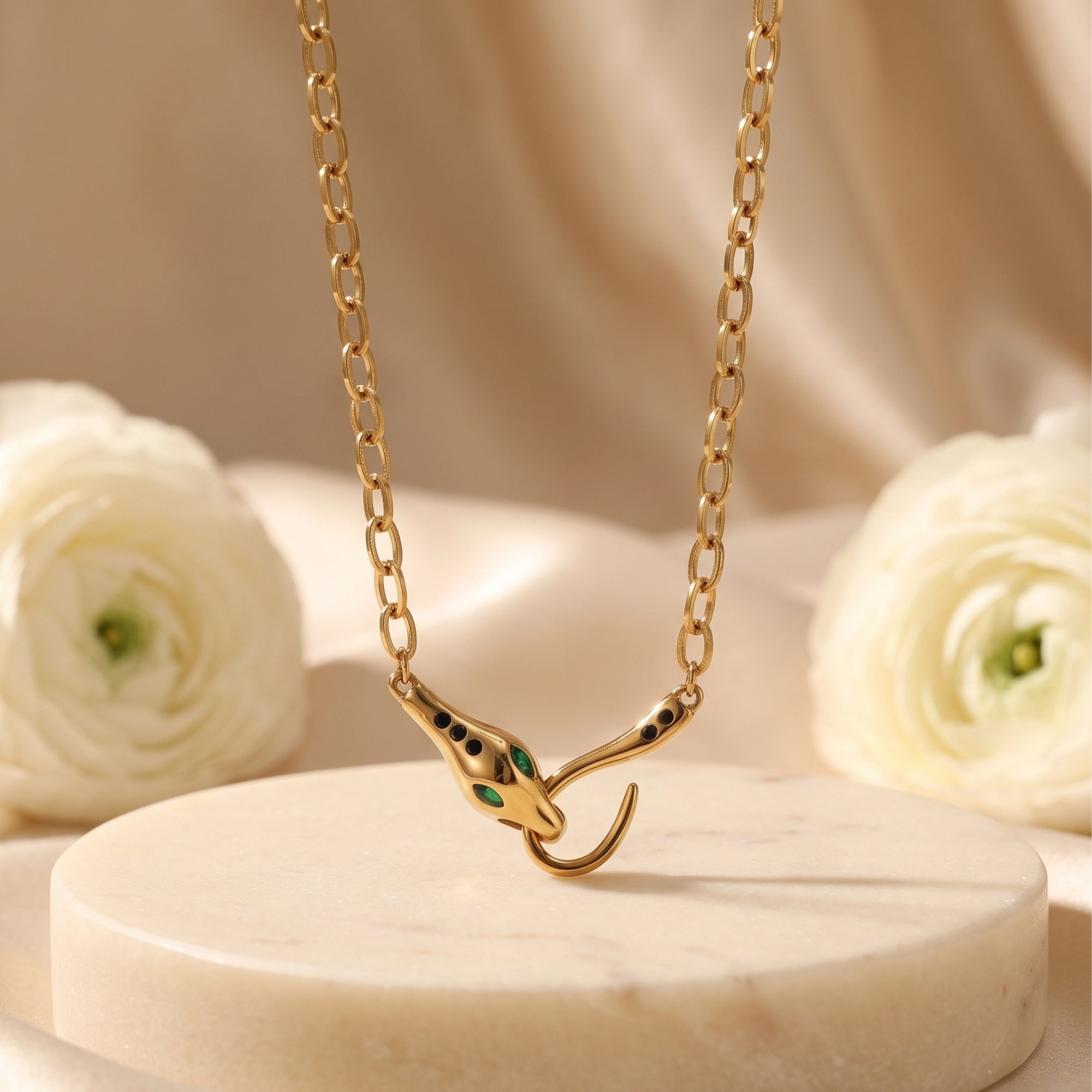 Gold Serpentine Necklace with a snake head clasp featuring green eyes, styled on a marble pedestal with flowers.