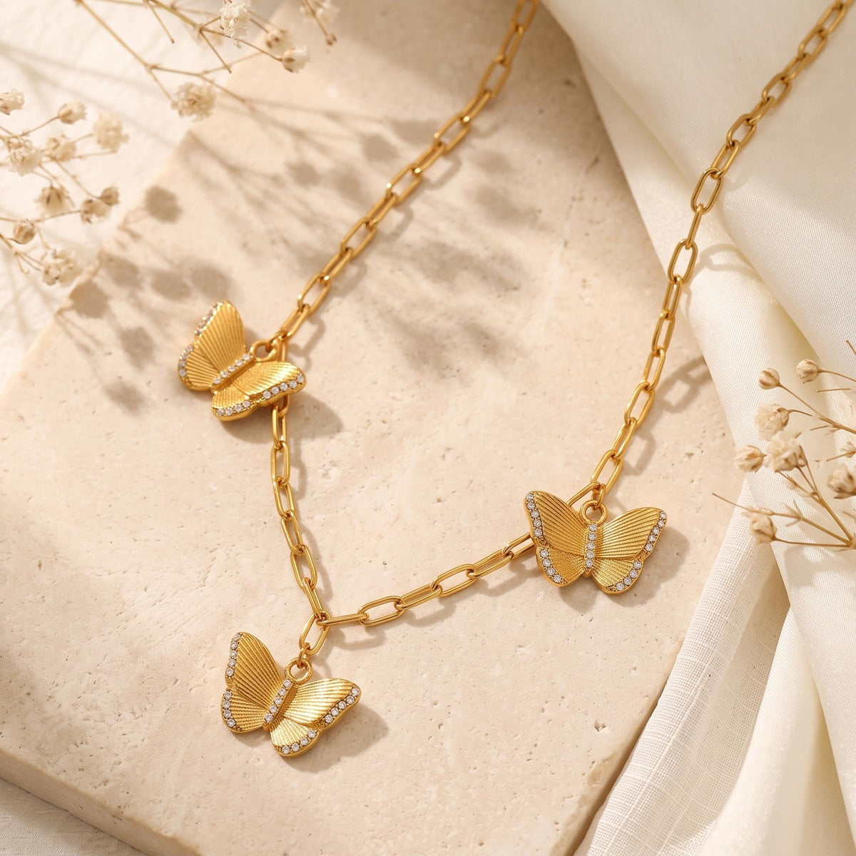 Gold-tone butterfly station necklace laid flat on textured stone next to white draped silk.