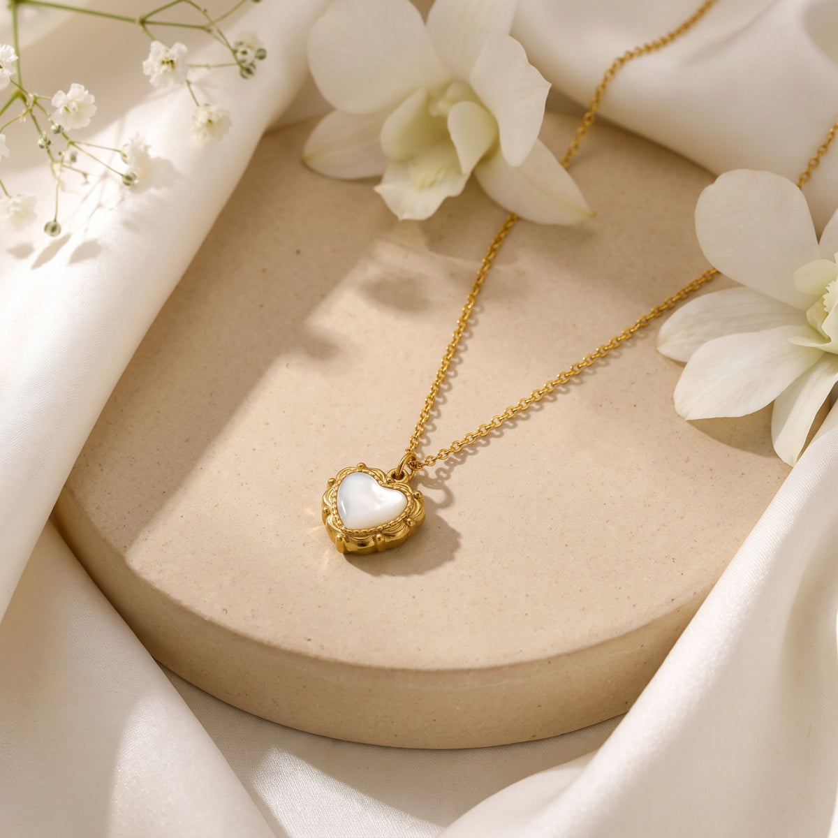 Gold-tone heart pendant necklace with a white mother-of-pearl simulant inlay on a round display platform with flowers.