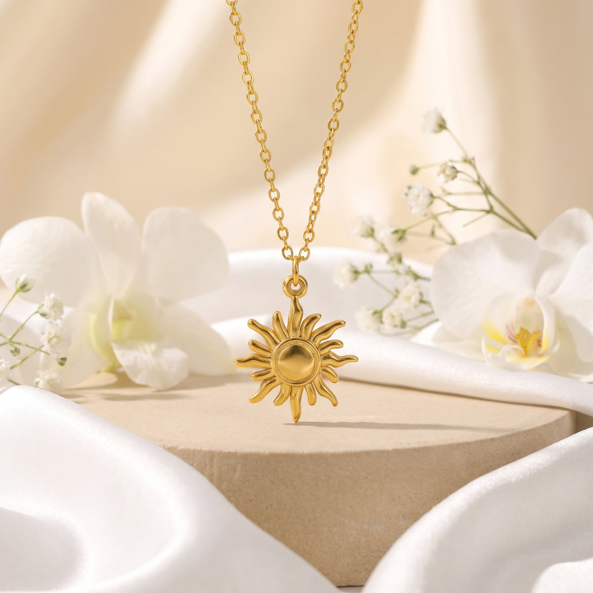 Gold sunburst pendant necklace displayed hanging above a beige pedestal with white orchids and draped silk.