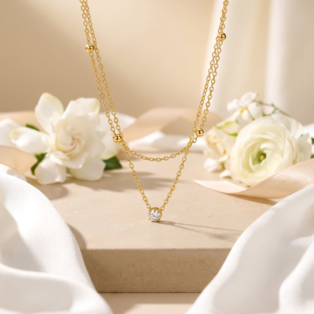 Gold-tone layered necklace with a beaded upper strand and clear solitaire pendant displayed among silk and white flowers.