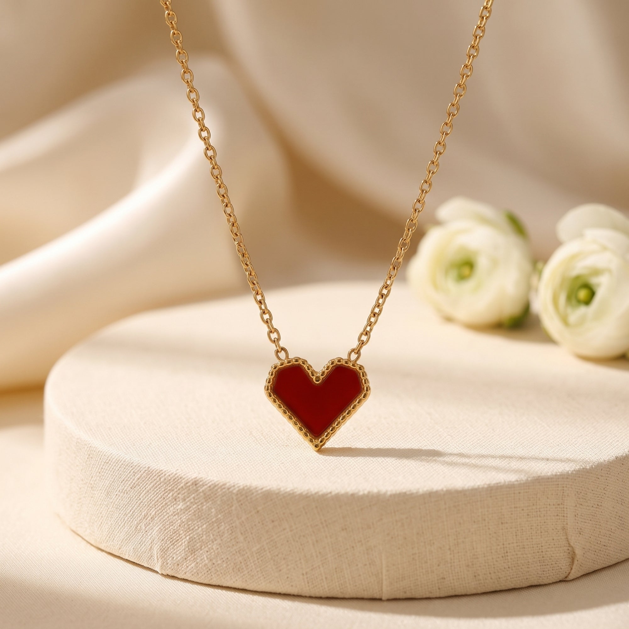 Scarlet heart necklace in 18K gold PVD stainless steel on a neutral pedestal with floral accents.