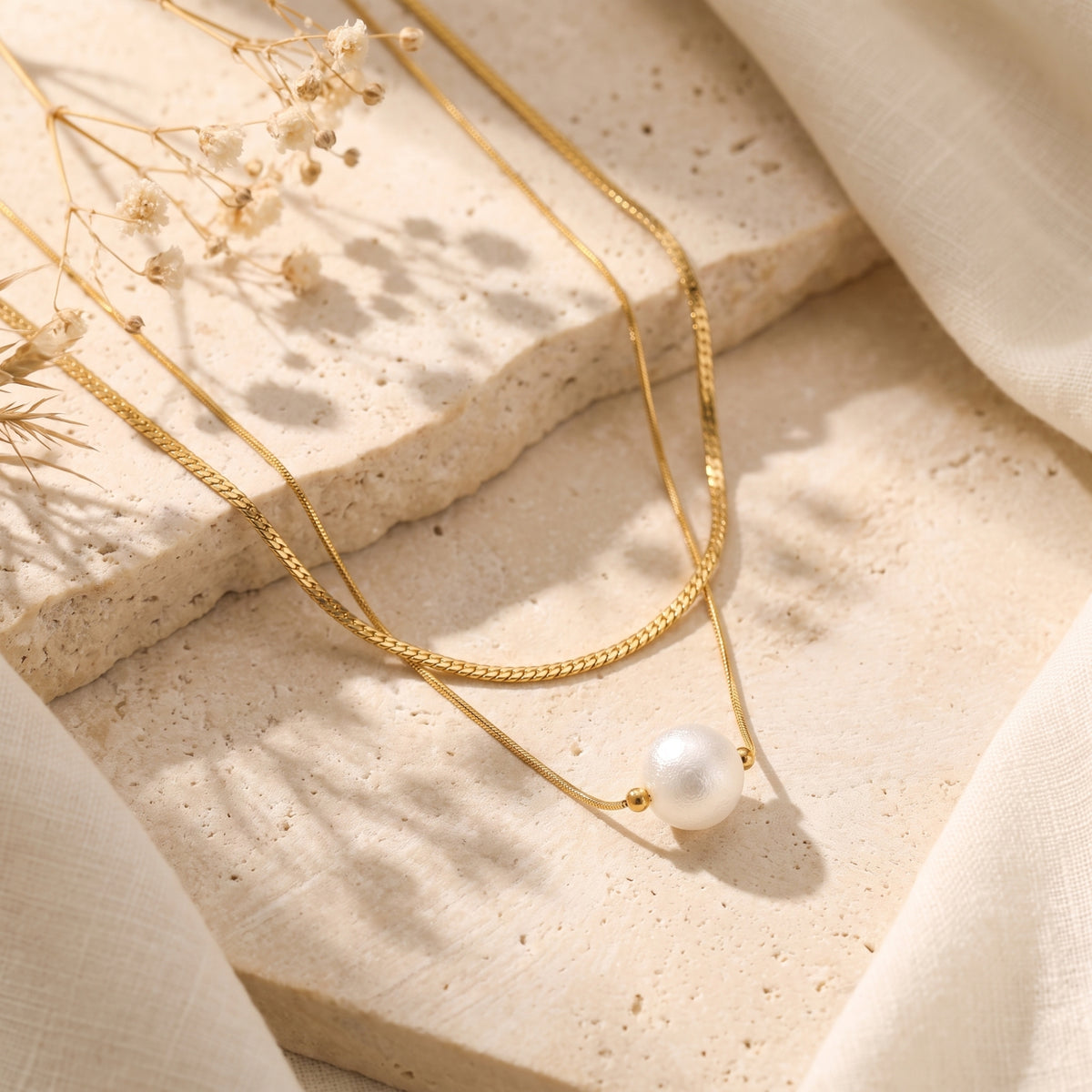 Layered gold-tone necklace with a white pearl pendant arranged on a pale textured stone surface with dried flowers.