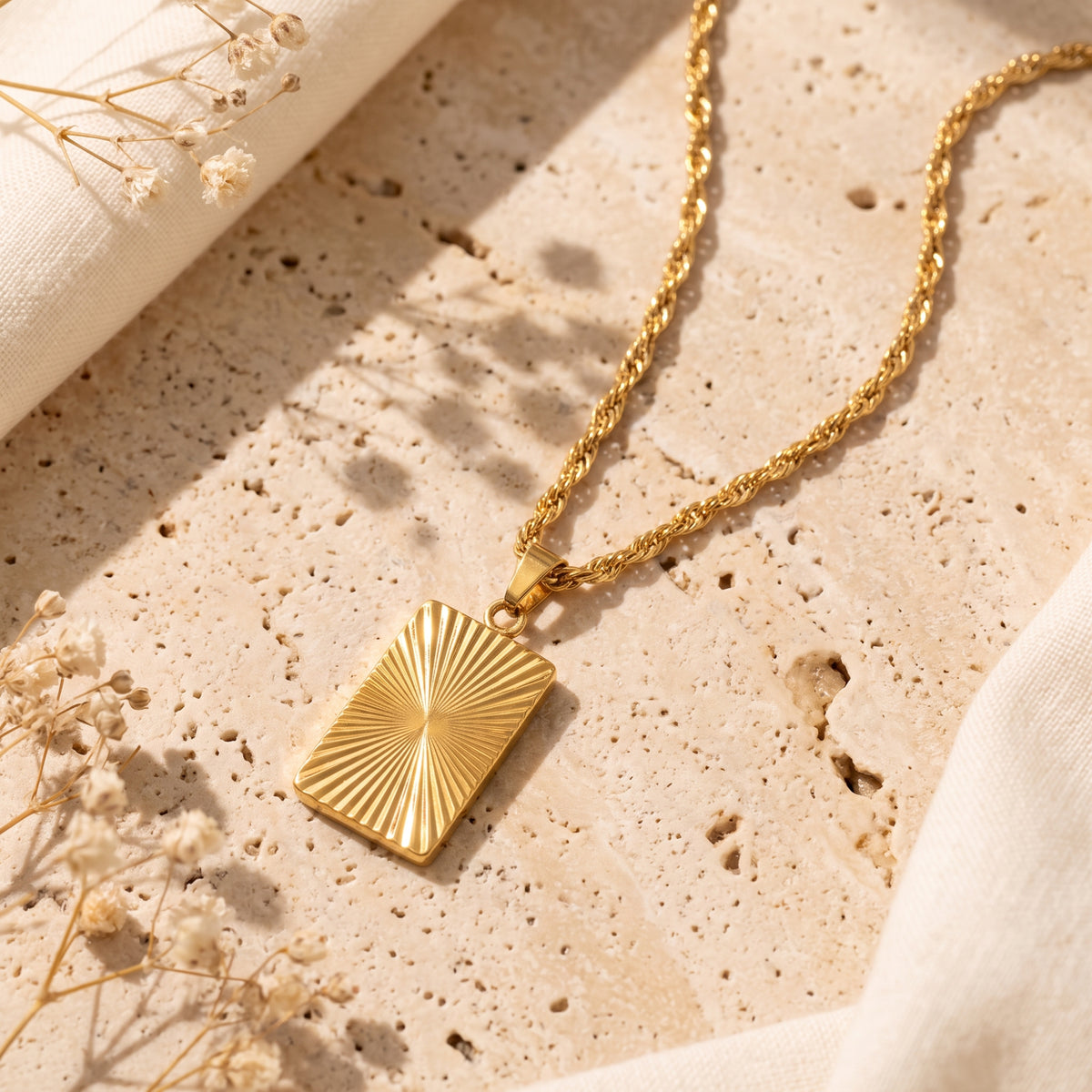 Gold-tone sunburst rectangular pendant on a twisted chain, lying flat on a beige textured slab with dried flowers.