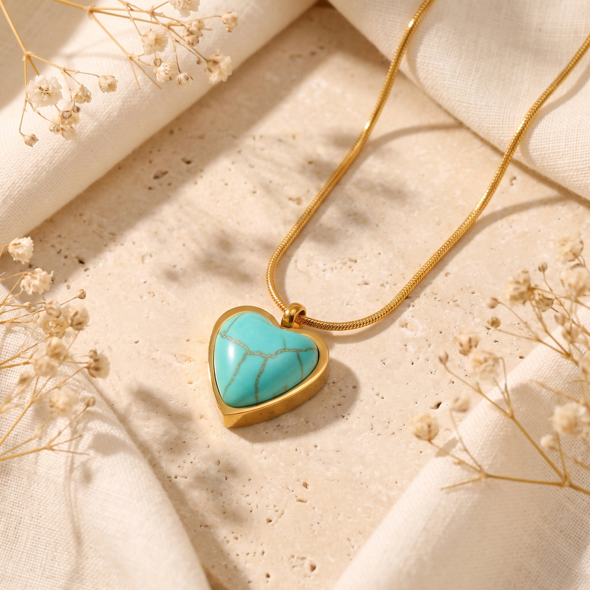 Flat lay of a gold-tone heart pendant with a turquoise stone on a beige stone slab among dried flowers.
