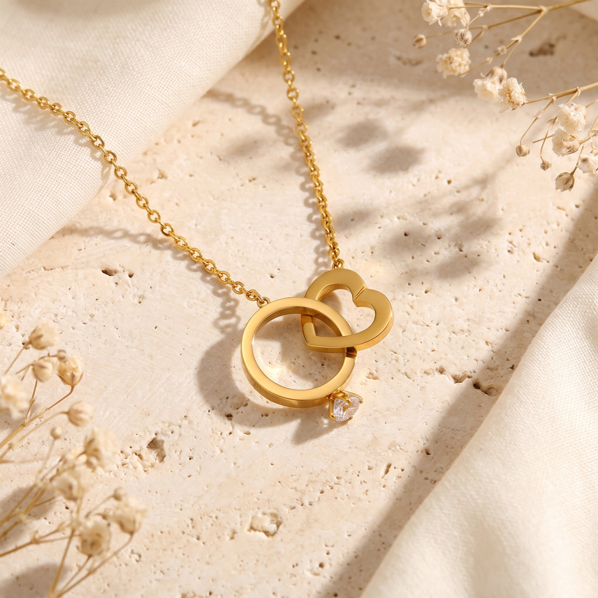 Gold-tone interlocking ring and heart necklace with a cubic zirconia accent, arranged on a beige stone surface.
