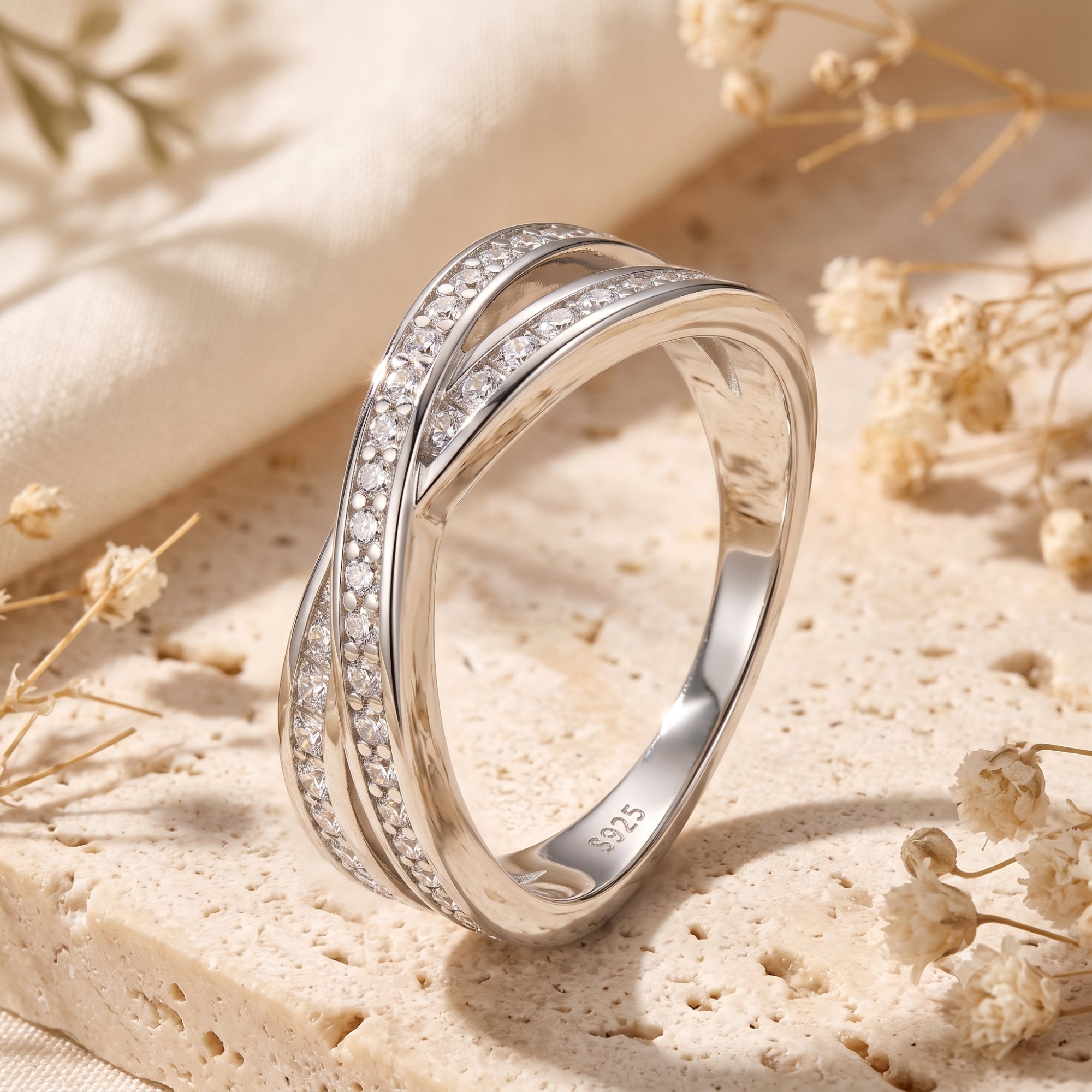 Sterling silver crossover ring with round simulated diamonds on a stone surface with dried flowers.