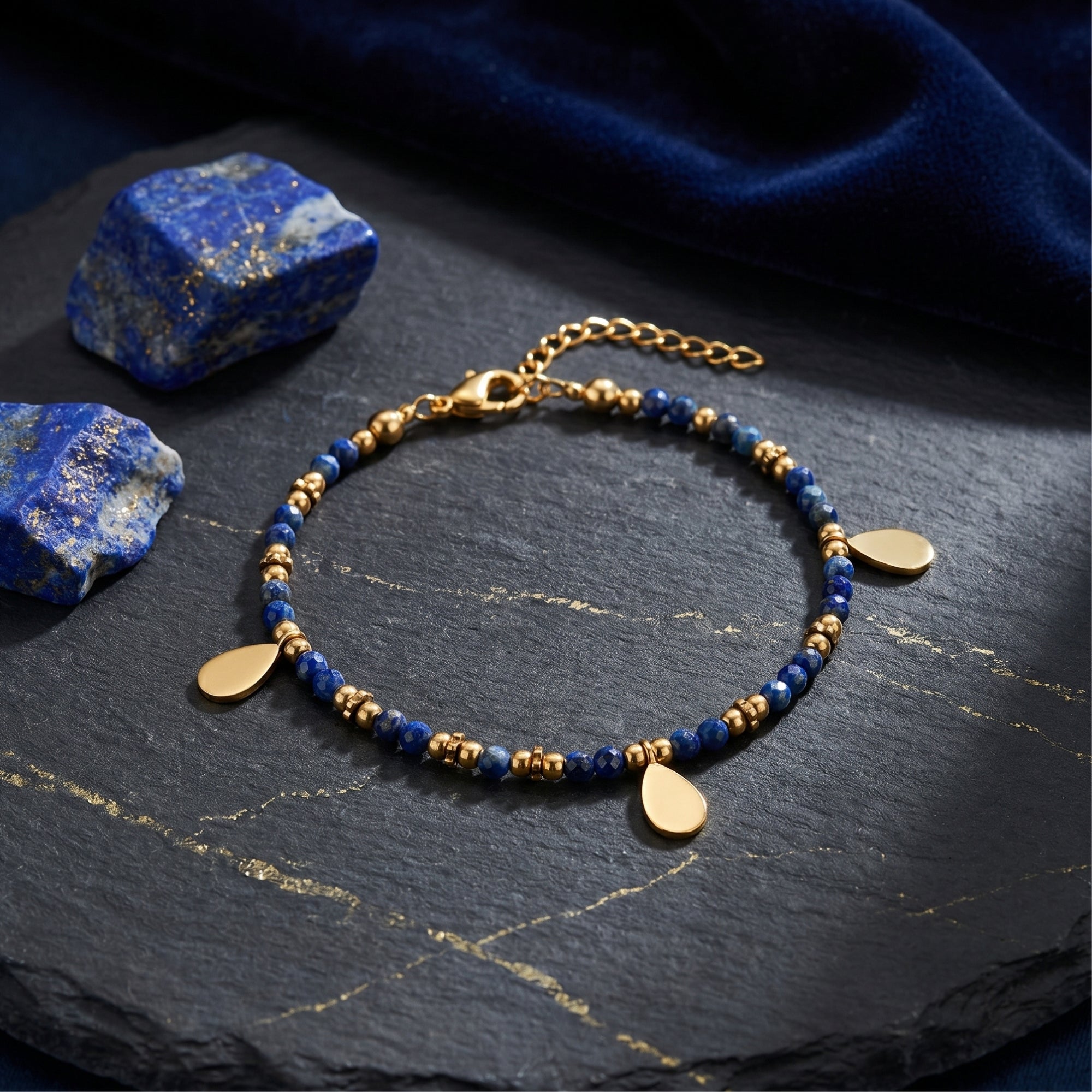 Gold plated stainless steel faceted 3mm lapis bead bracelet with teardrop charms on slate with raw stones.