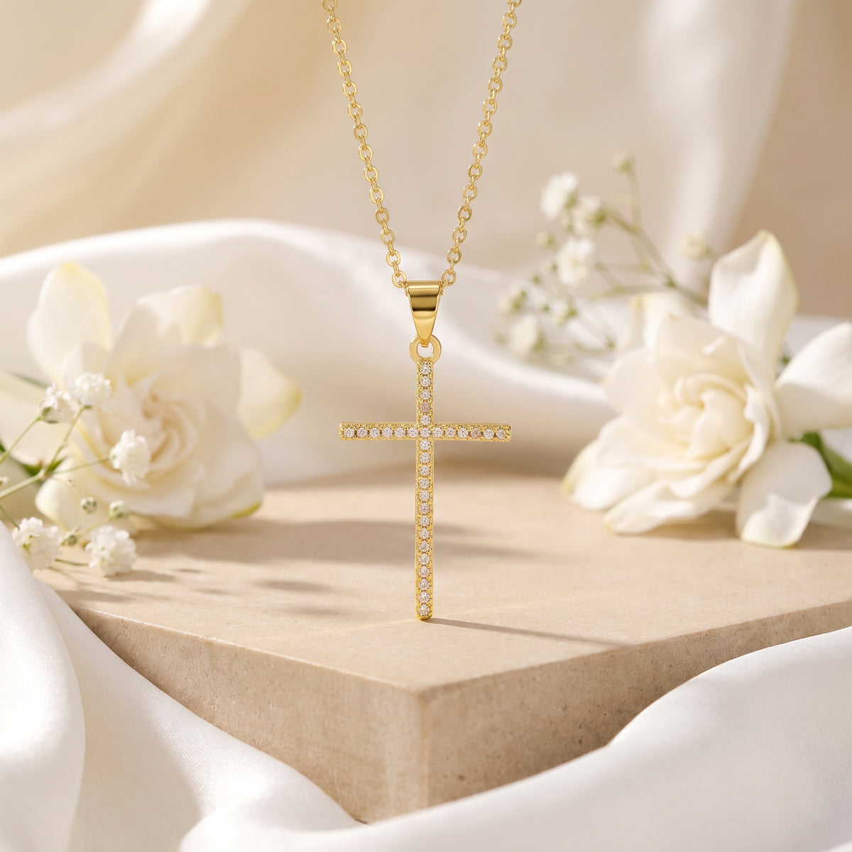 Gold-tone cross pendant set with clear pavé stones hanging above a beige block with white silk and flowers.