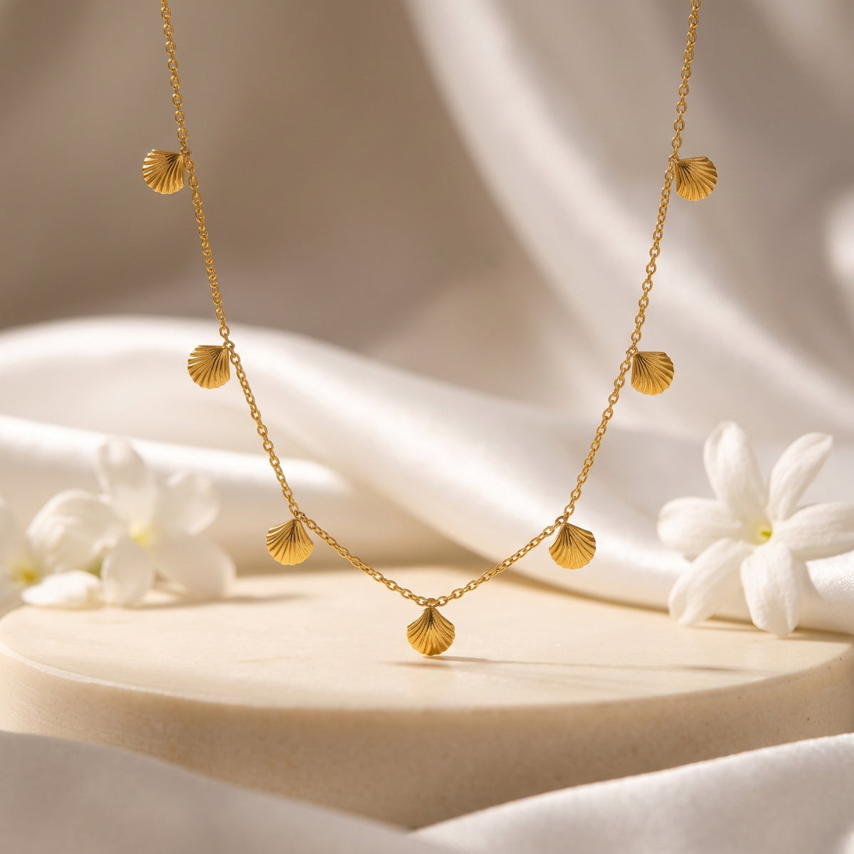 Gold-tone scallop shell station necklace suspended above a round beige pedestal with white flowers.