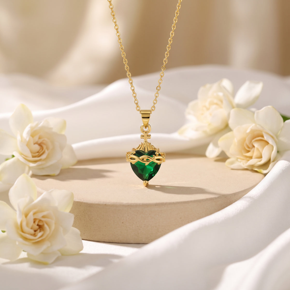 Gold-tone necklace with a heart-cut simulated emerald pendant, cross, and crown-of-thorns detail hanging above white flowers.