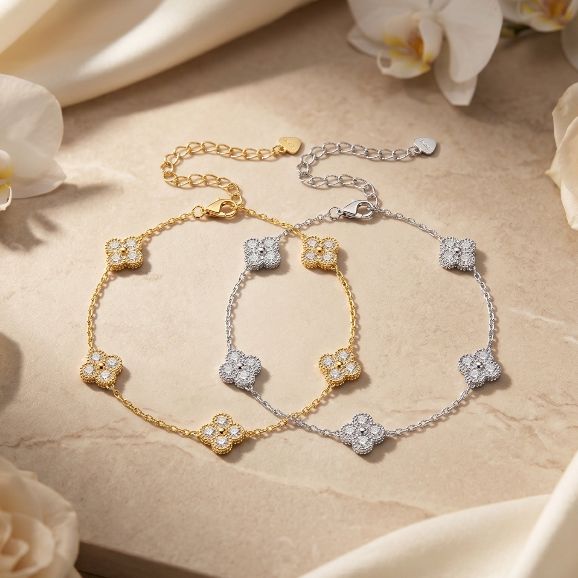 Gold and silver clover bracelets with moissanite stones on a stone surface with white flowers.