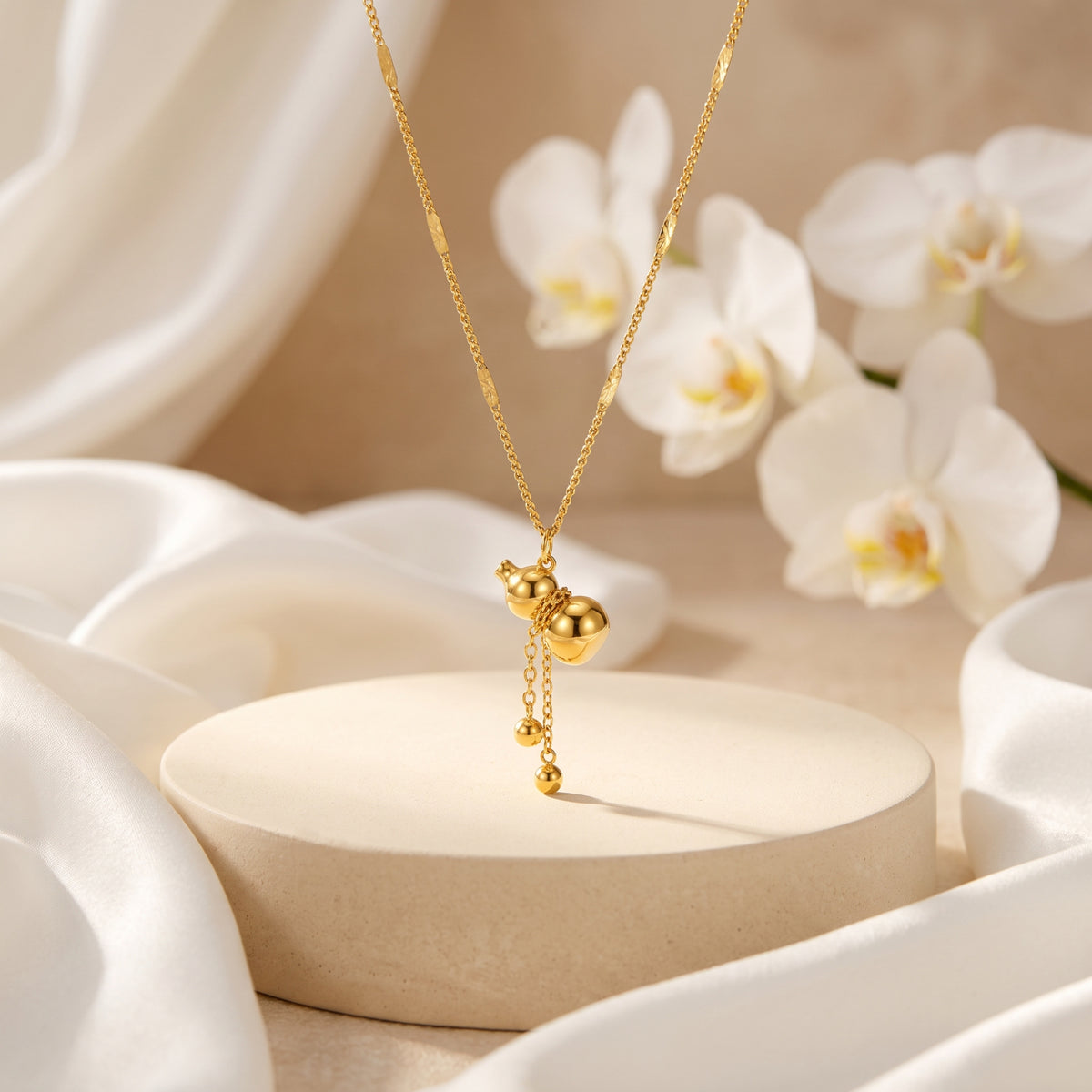 Gold-tone gourd drop necklace suspended above a round beige pedestal with draped white silk and orchids.