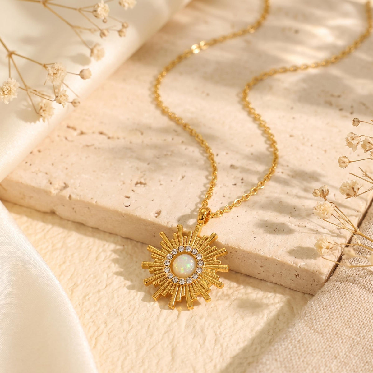 Gold-tone sunburst opal pendant necklace laid on a beige stone tile beside white fabric and dried flowers.