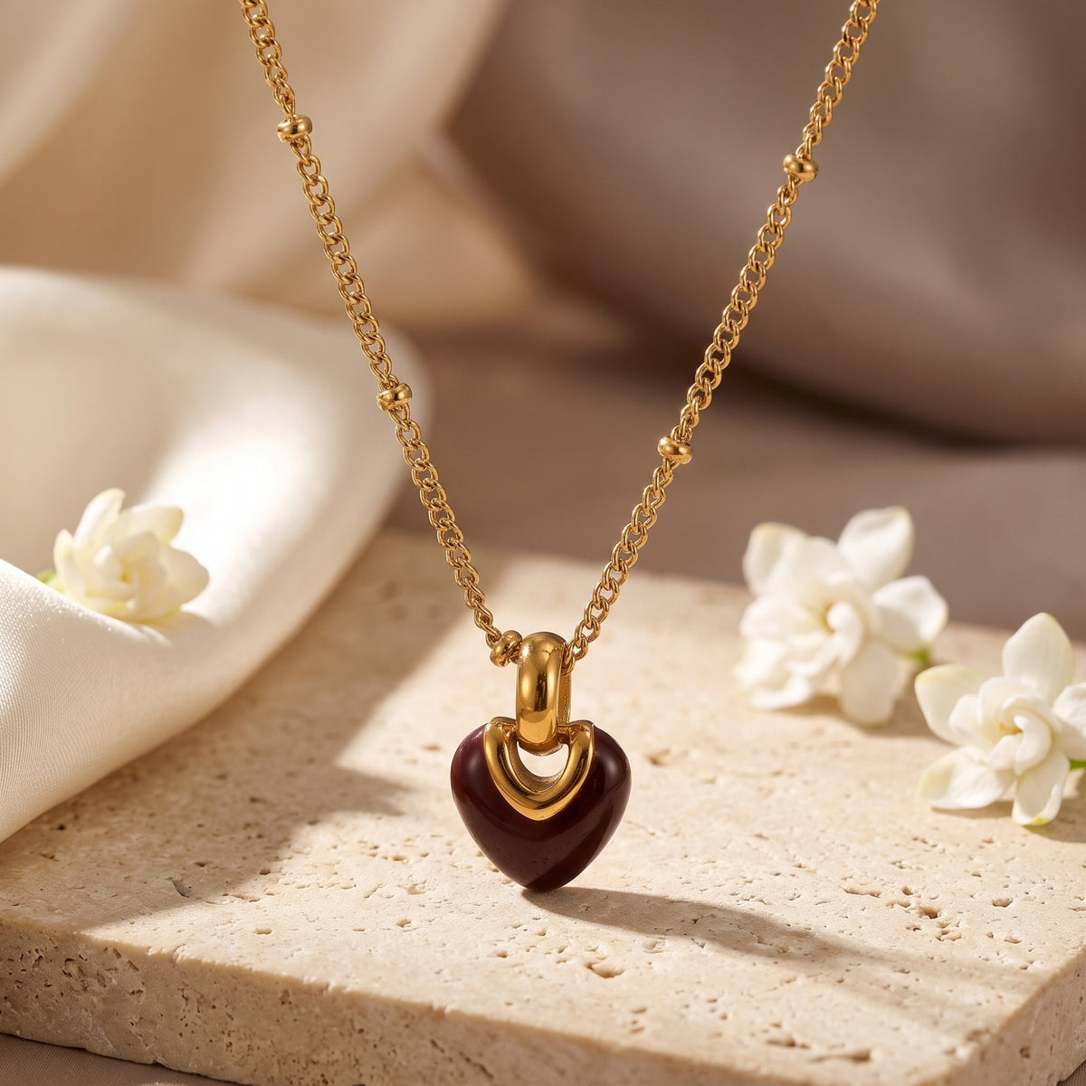 Front close-up of a gold-tone beaded chain necklace with a glossy crimson heart pendant on a stone block.