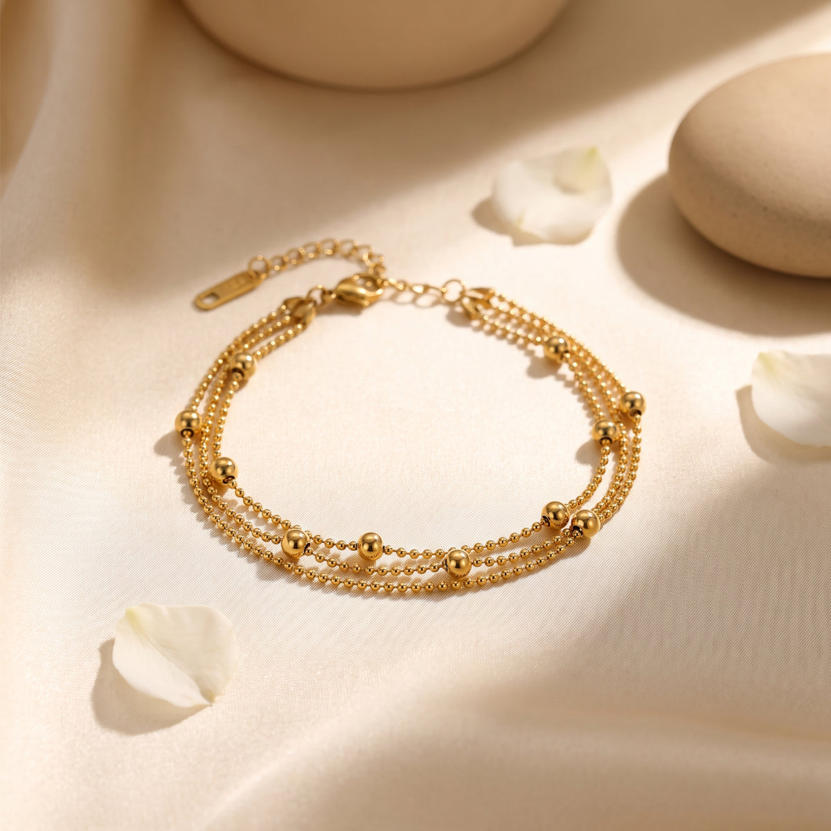 Gold-tone three-strand bead bracelet arranged in a loop on draped fabric with scattered white petals.