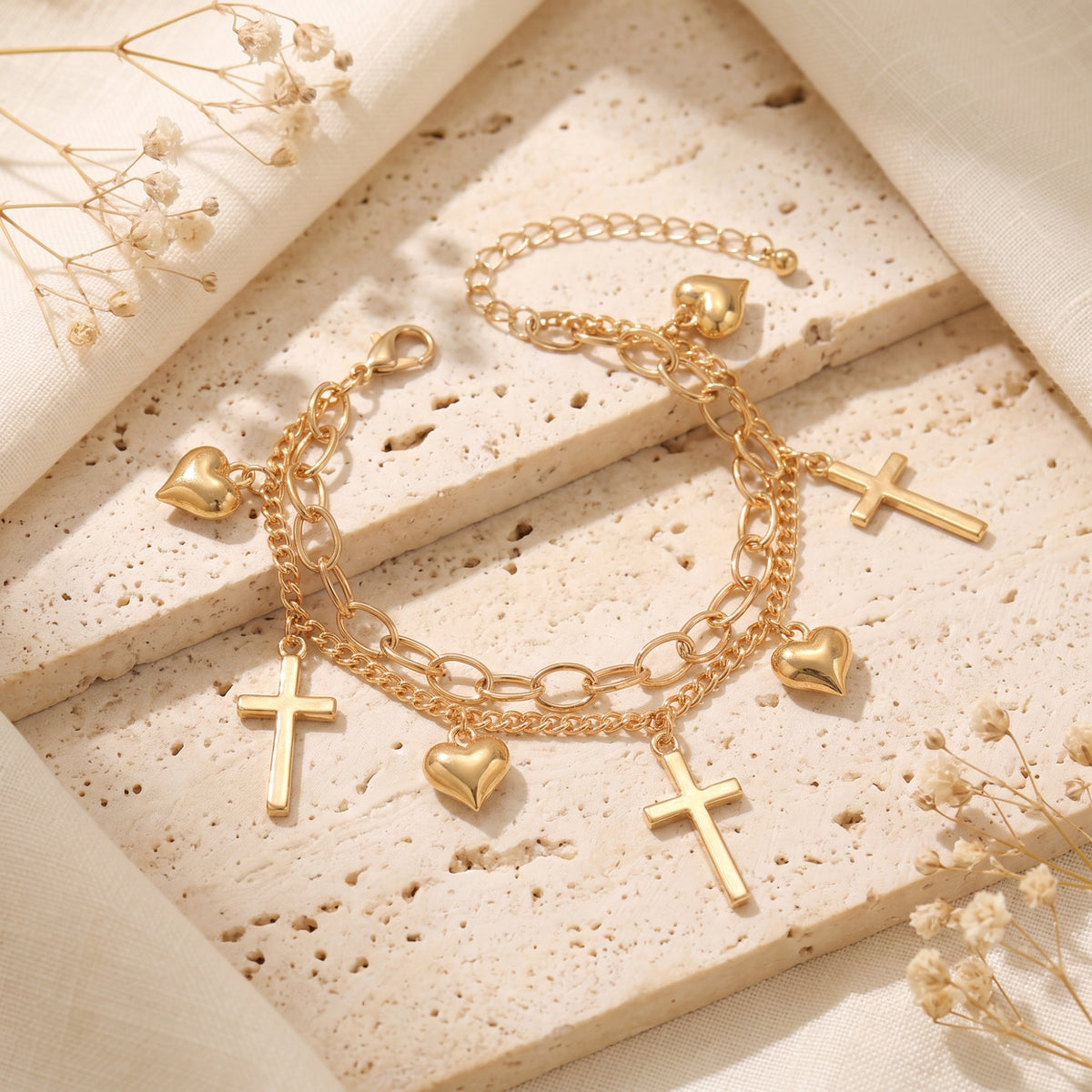Gold-tone double-chain charm bracelet with heart and cross charms lying open on a textured beige stone surface.