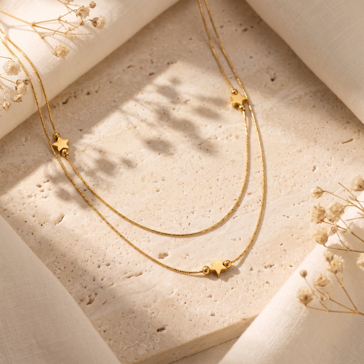 Double-strand gold-tone star necklace lying on a stone tray among linen fabric and dried flowers.