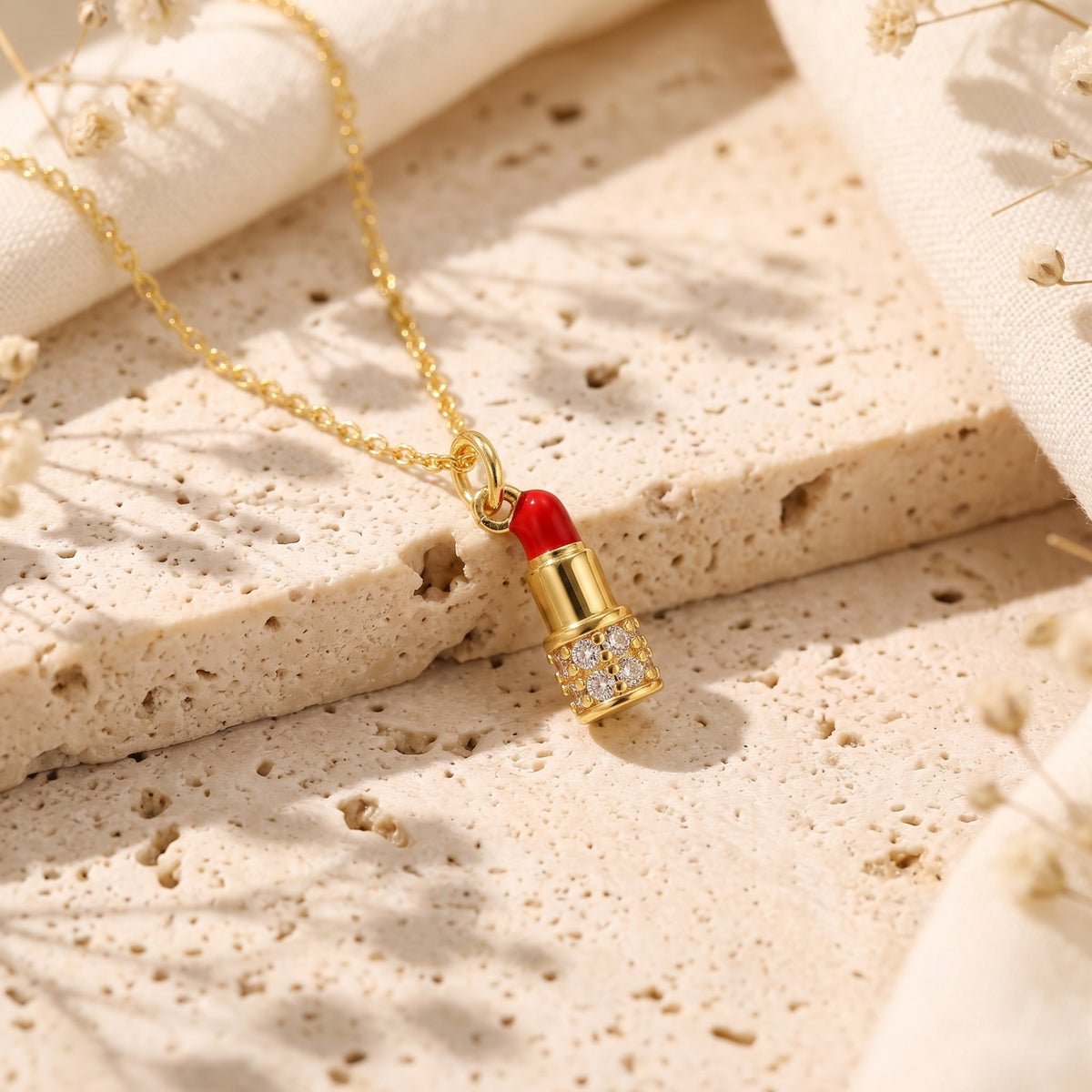 Gold-tone lipstick pendant with a red enamel tip and simulated diamond accents on a porous stone surface.