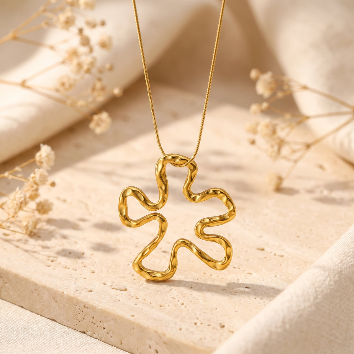 Gold-tone openwork wavy flower pendant hanging from a fine chain over a beige stone slab with dried flowers.