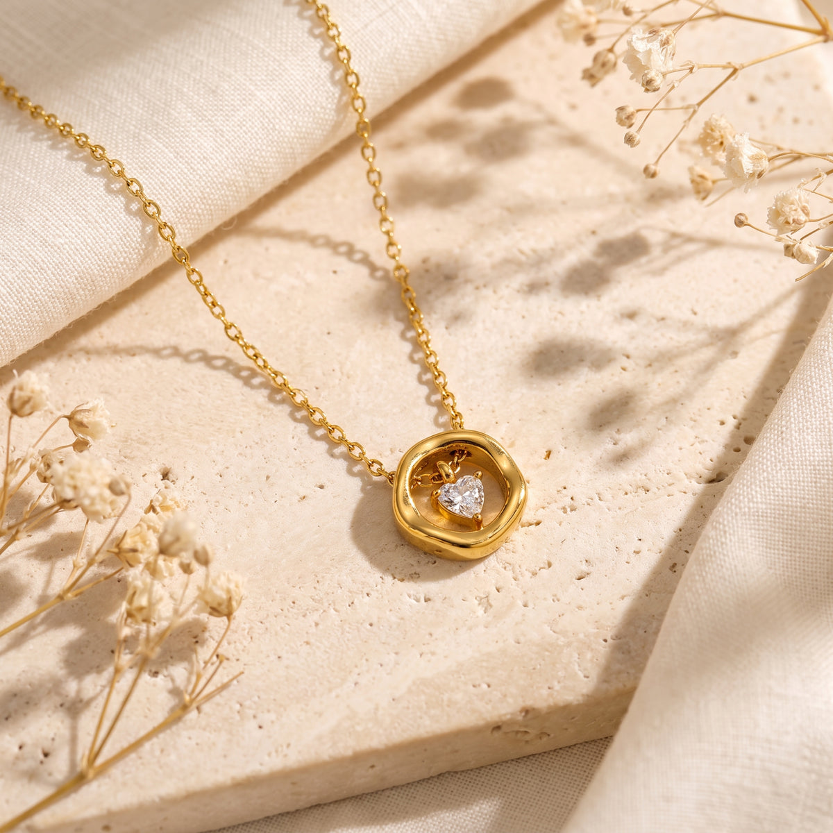 Gold-tone necklace with a heart-shaped clear stone inside a rounded ring, arranged on a stone slab with dried flowers.