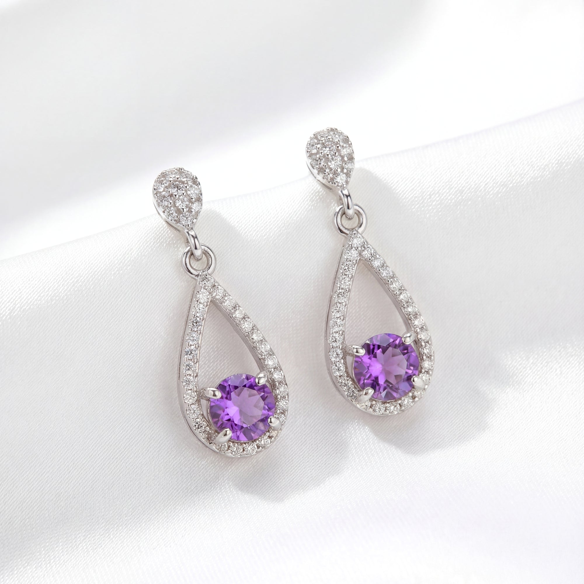 Sterling silver teardrop halo drop earrings with round simulated amethyst on white silk fabric.