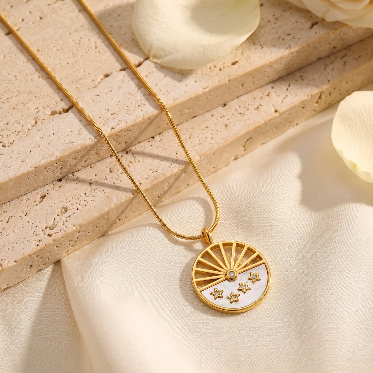 Gold-tone celestial sunburst pendant with star accents resting on textured stone with cream petals.