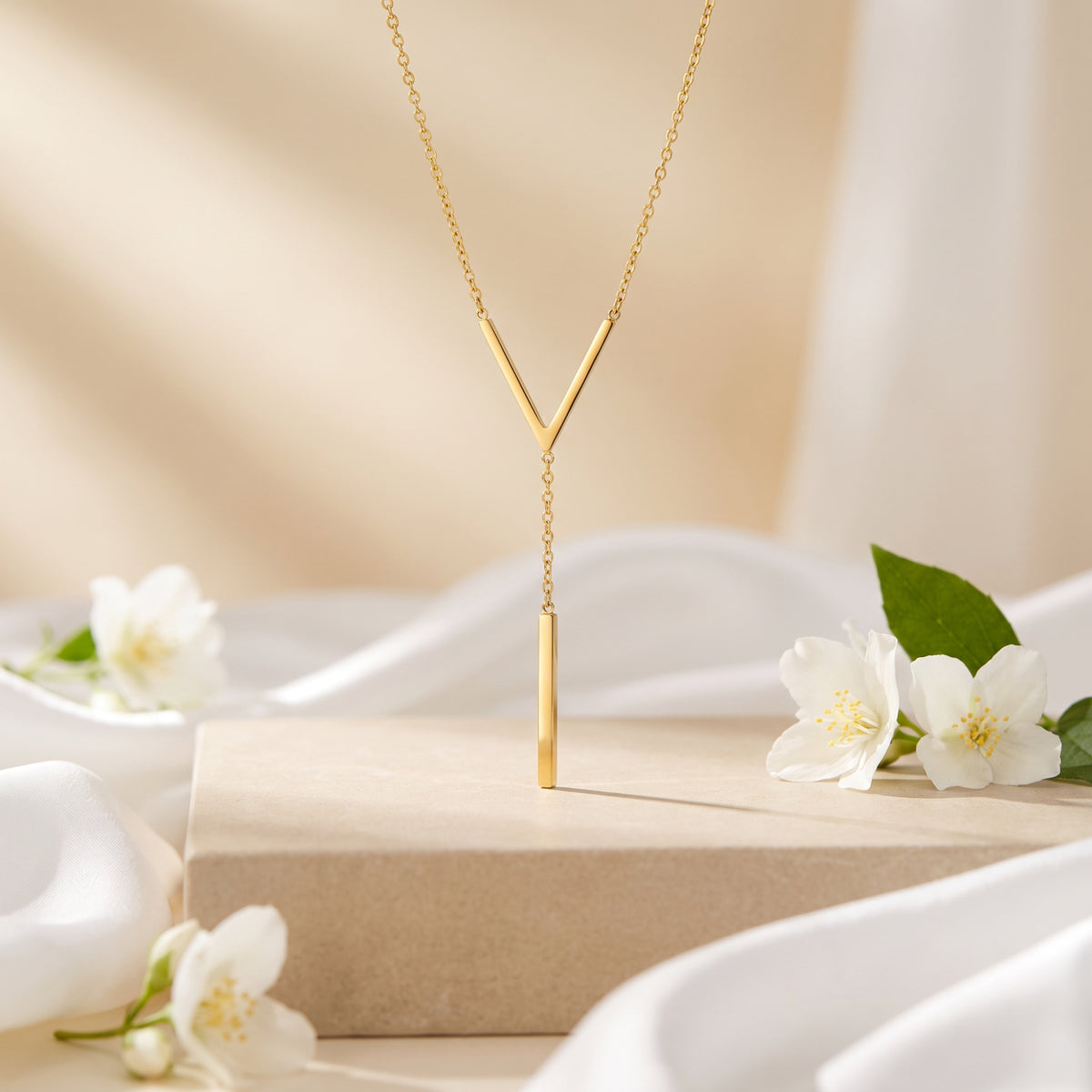 Gold-tone chevron lariat necklace hanging from a beige pedestal beside white flowers.
