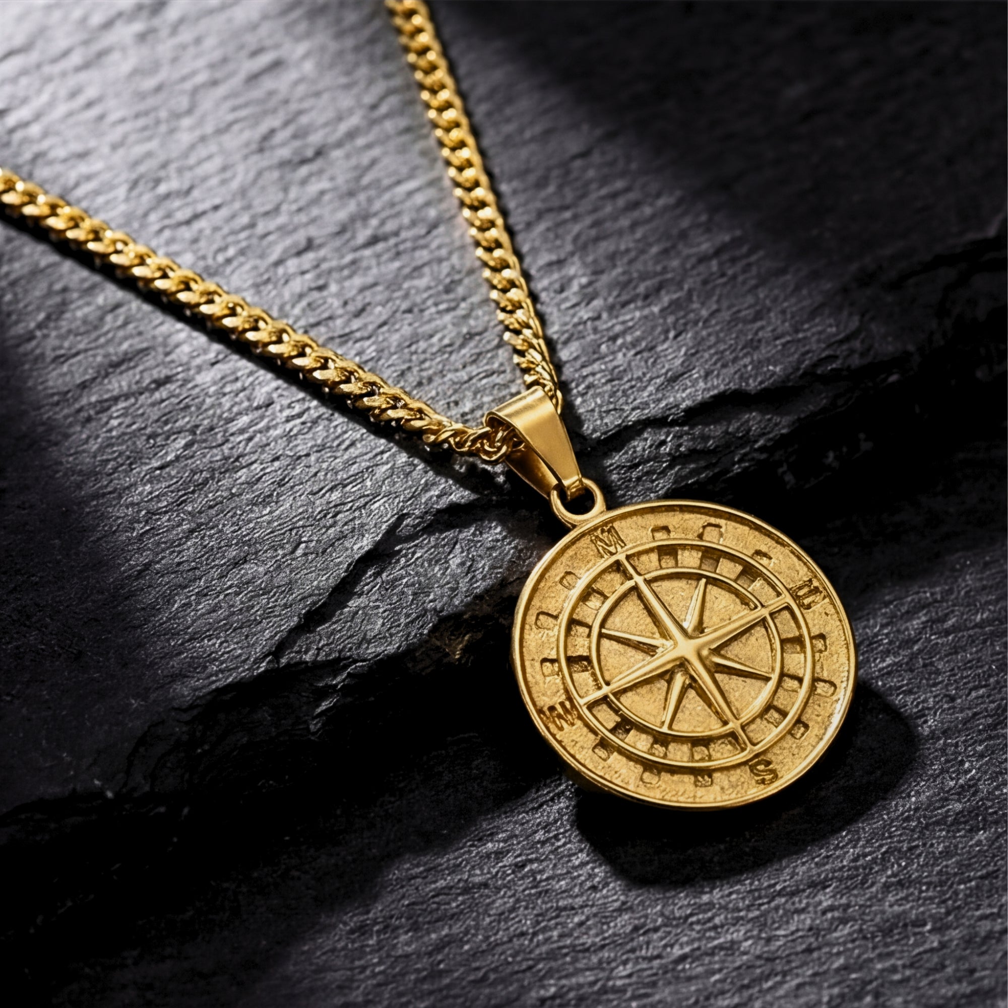 Gold stainless steel Voyager compass pendant on a curb chain, resting on a dark textured stone surface.