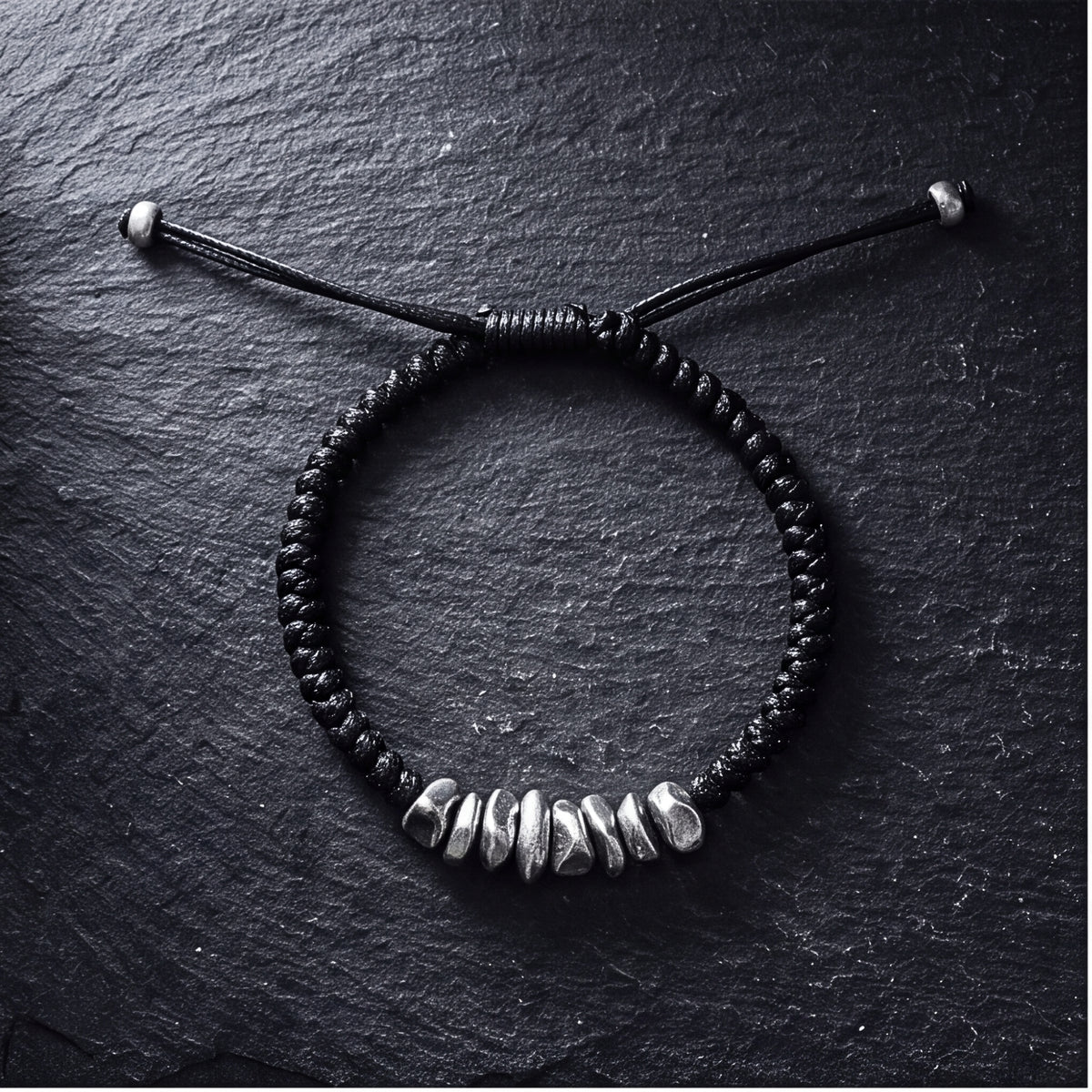 Black braided cord bracelet with silver nuggets arranged in a circle on dark slate.