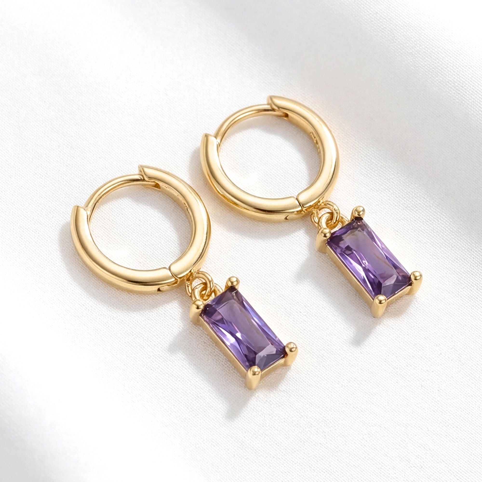 Gold hoop earrings with emerald cut simulated amethyst drops resting on white textured fabric.