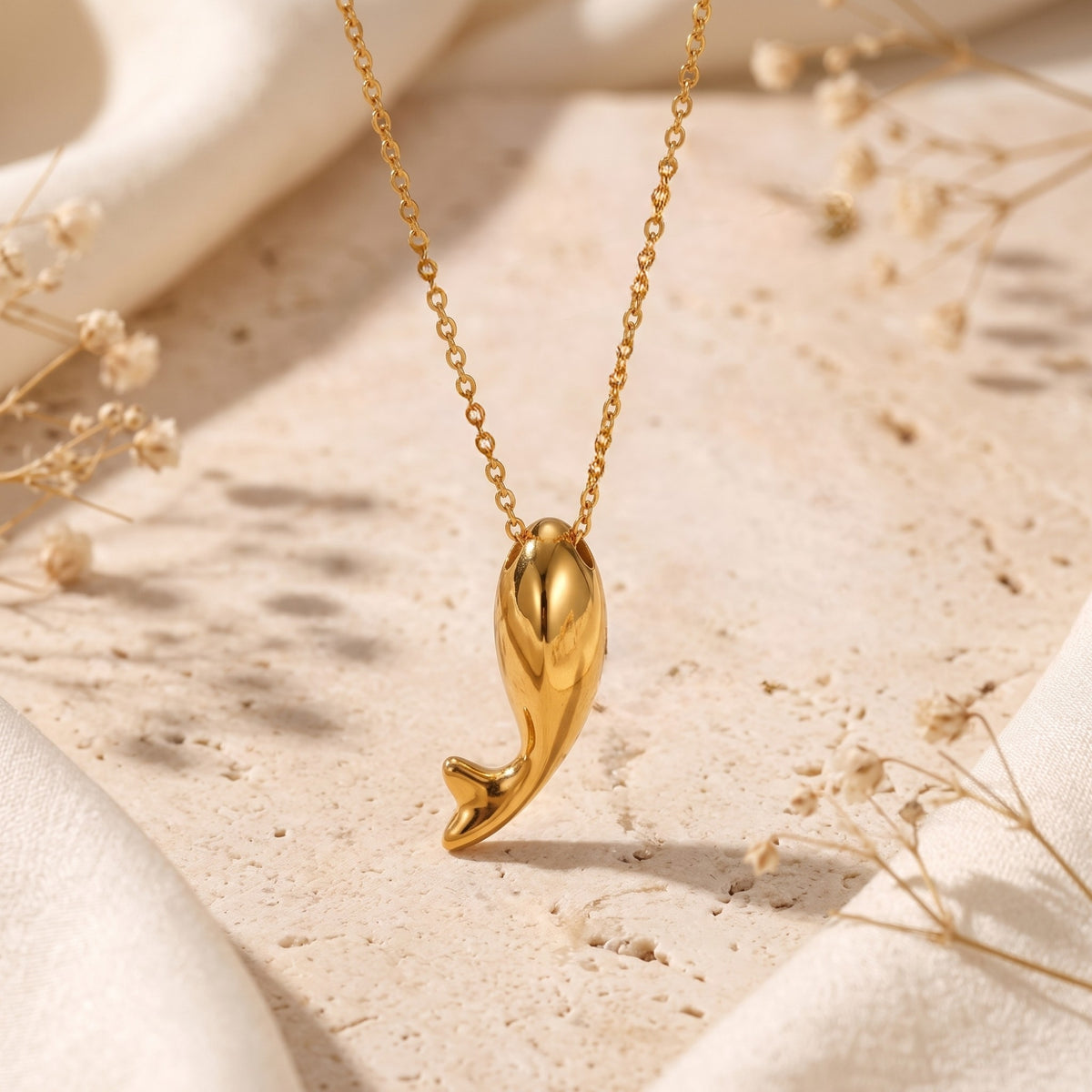 Smooth gold-tone dolphin tail pendant suspended vertically on a delicate chain against a stone background.