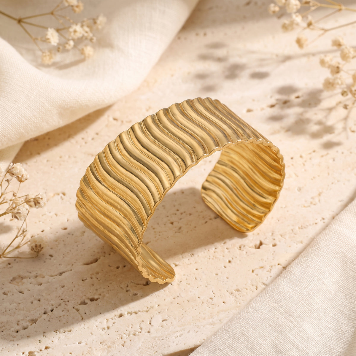 Gold-tone cuff bracelet with sculpted wave ridges displayed on a stone surface.