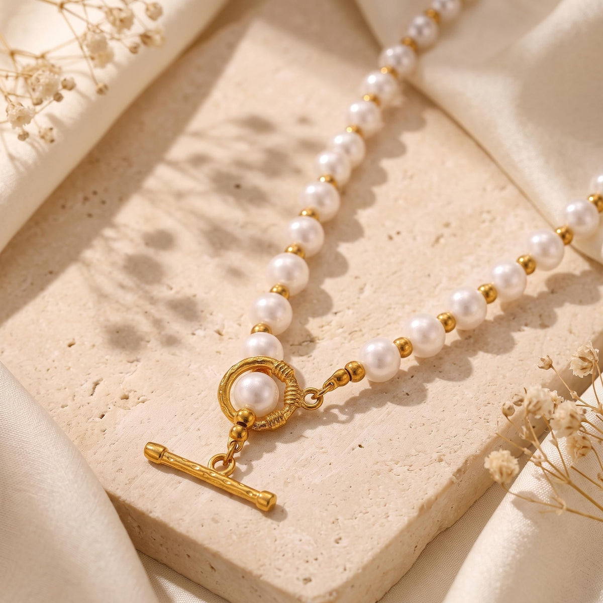 Close-up of a white simulated pearl necklace with gold-tone spacer beads and a T-bar toggle clasp on a stone slab.
