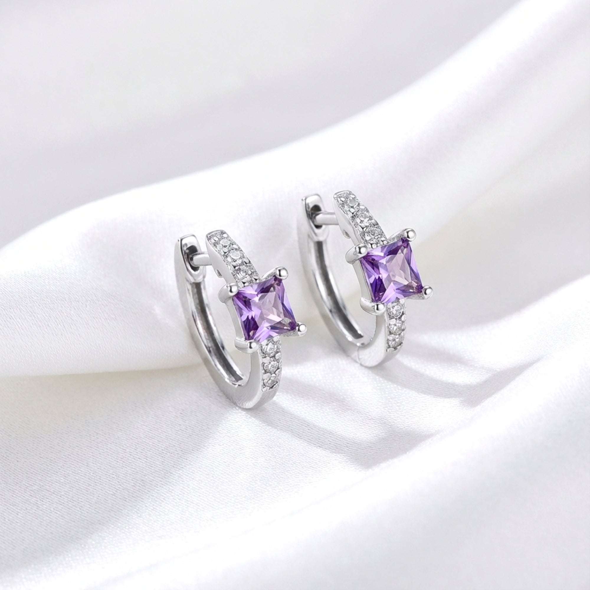 Sterling silver huggie earrings with princess-cut simulated amethyst and clear accents on white silk.