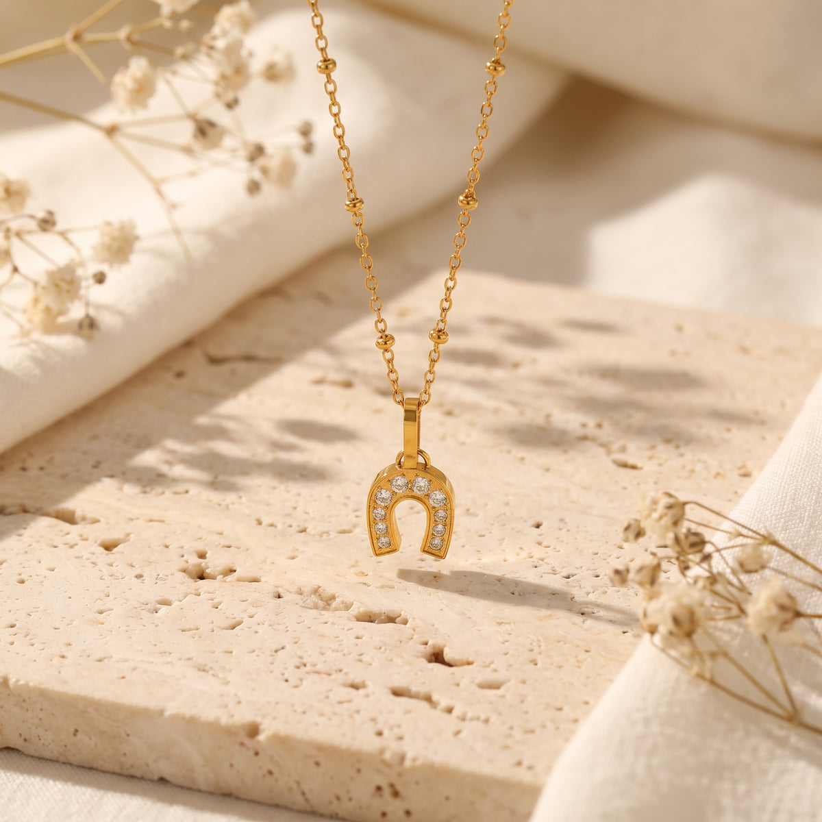 Gold-tone horseshoe pendant with clear stones on a bead-station chain, hanging above a textured stone slab.